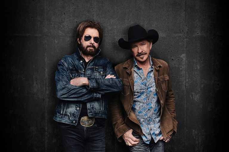 Country music duo Brooks & Dunn pose against a dark wall; Kix Brooks wears a cowboy hat and Ronnie Dunn wears sunglasses in a denim jacket.